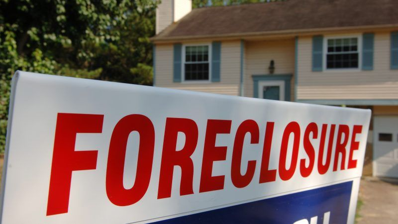 A large "foreclosure" sign in red letters, prominently displayed in front of a two-story suburban house under a clear sky.