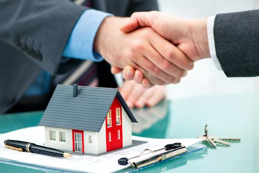 Two individuals shaking hands over a desk with a small model house, keys, and paperwork, indicating a real estate transaction or agreement.