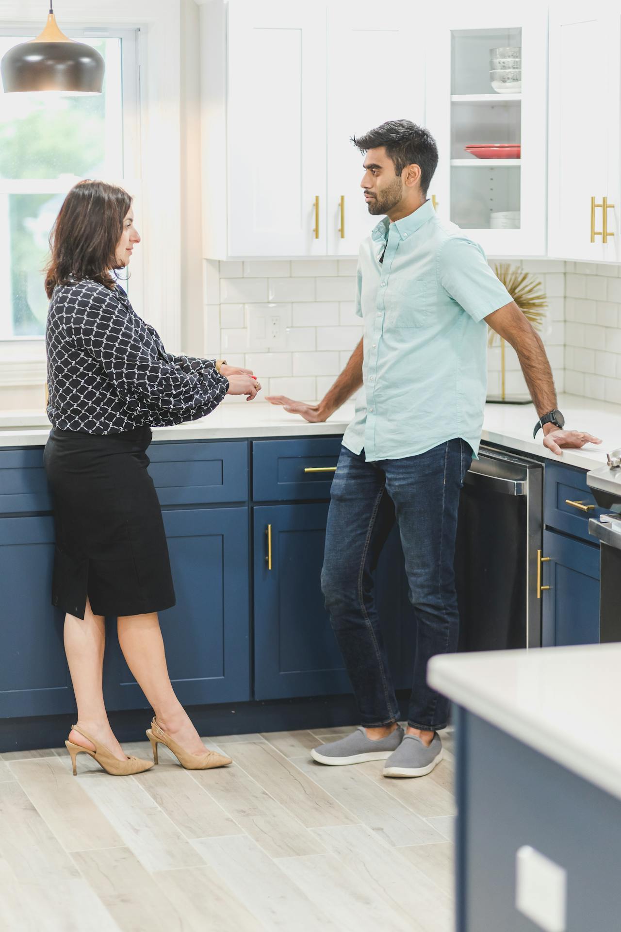 A woman dressed in a patterned blouse, black skirt, and beige heels stands in a kitchen facing a man in a pale blue button-down shirt and jeans. They appear to be engaged in a conversation. The kitchen has blue lower cabinets and white upper cabinets.